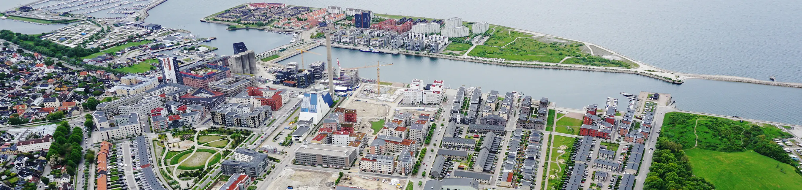 Flygfoto över en urban kustmiljö med bostadshus och parker vid vattnet. Bilden visar en tät stadsbebyggelse med färgglada byggnader, gångvägar, grönområden och en marina i bakgrunden. 