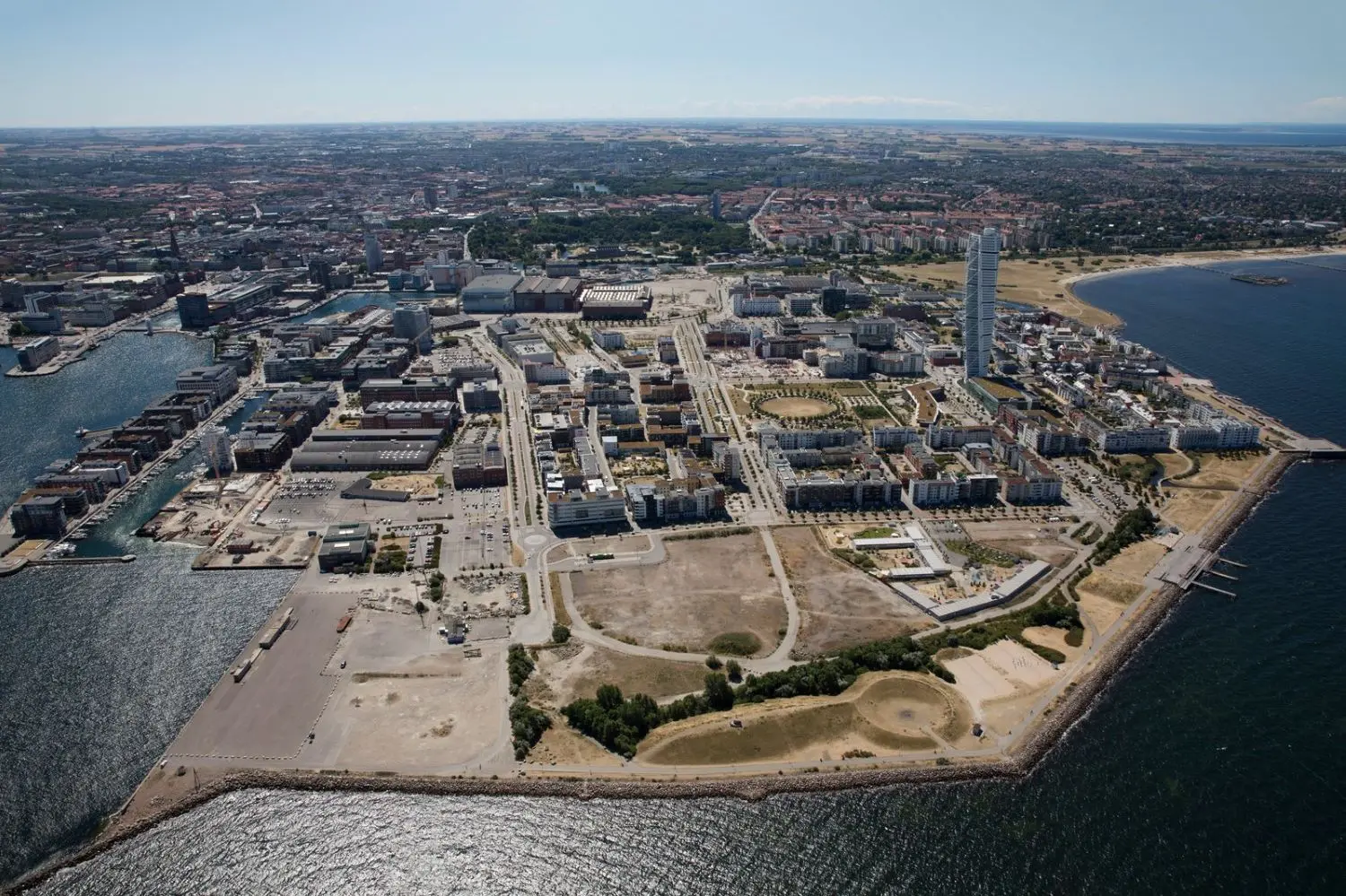 Flygbild över Västra Hamnen i Malmö med Turning Torso som sticker upp bland byggnaderna och stadens kvarter utspridda längs kustlinjen. Vattnet omger området, och parker och öppna ytor syns nära havet.