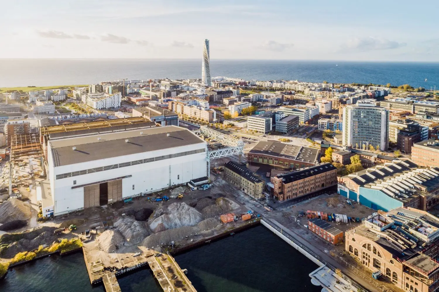 Bild över industriområdet i Malmö med byggnader i olika stadier av renovering och rivning, samt Turning Torso synlig i bakgrunden nära havet. Området omges av kanaler och vattnet syns i bildens nederkant