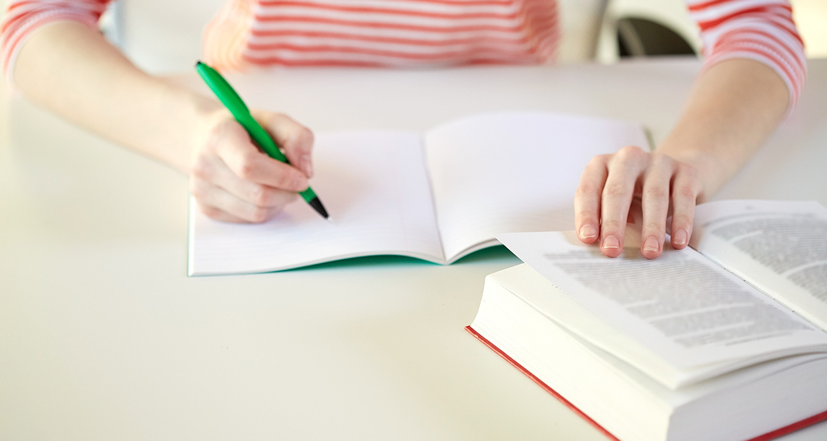 Ett skrivhäfte är uppslaget och man ser två händer. Den ena handen håller i en penna. Bredvid ligger en uppslagen bok. Personen har en röd- och vitrandig tröja på sig.