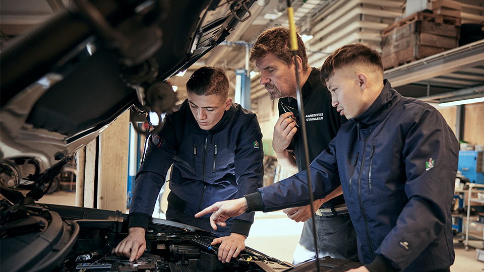 Två elever och en lärare kollar ner i en personbilsmotor.