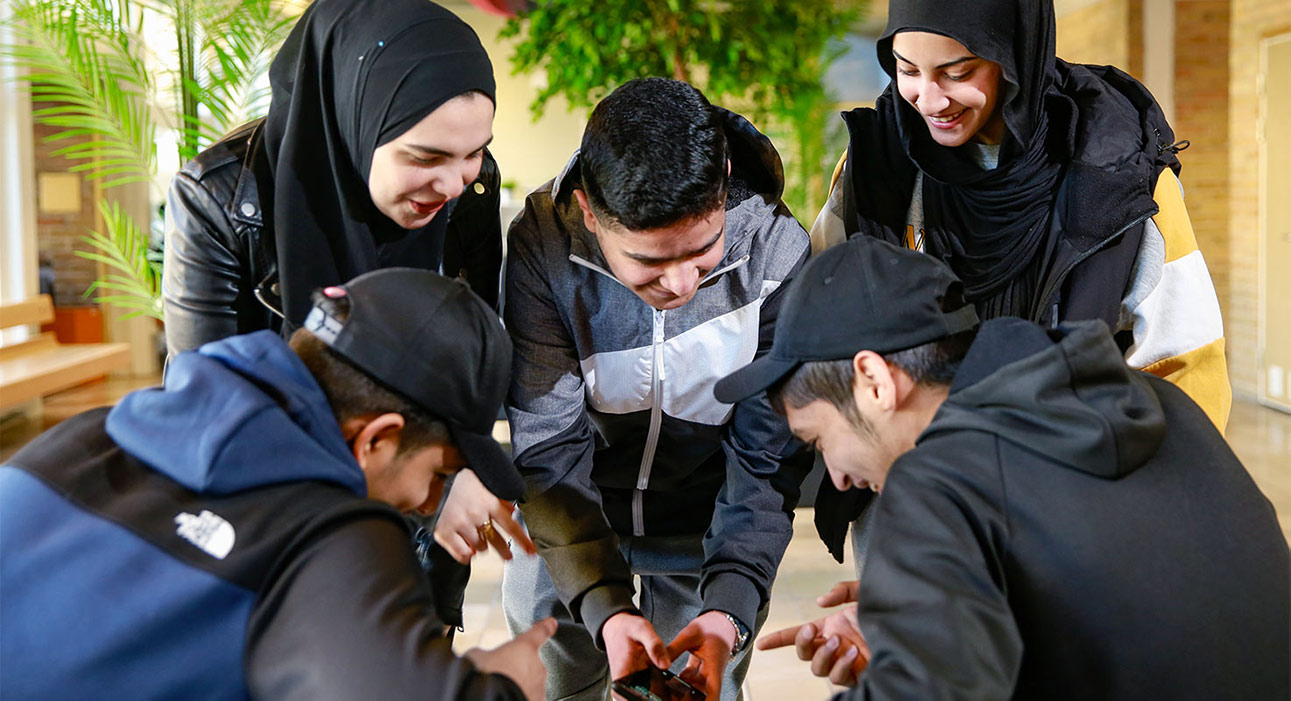 Fem glada elever samlade kring en mobiltelefon, skrattande och engagerade i något gemensamt. Positiv stämning och samarbete syns tydligt.