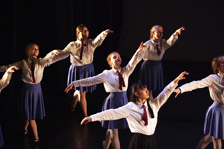Stämningsfull ljussättning på en scen med flera unga kvinnliga dansare i kjol, slips och vita skjortor. 