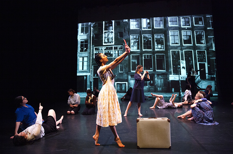 En scen med stämningsfull scenografi i form av stadsmiljö. Dansare som står och sitter för att skapa en känsla av ett levande torg.