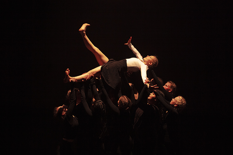 En dansare lyfts upp på scenen av andra dansare. Ljuset faller majestätiskt på dansaren som ligger på de andras händer. 