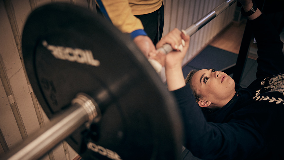 En elev tränar i en bänkpress i skolans gym. På bilden syns också en annan elevs händer som hjälper den tränande. 