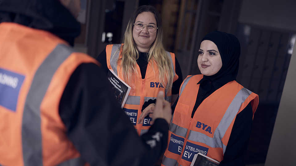 Två elever som läser till väktare står och samtalar med en tredje elev. Eleverna bär orange reflexväst med texten säkerhetsutbildning elev.
