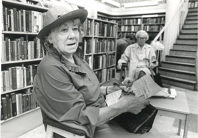 Porträttfotografi på Herta Wirén. Hon håller ett tidningsurklipp i handen och sitter i ett rum med många bokhyllor.