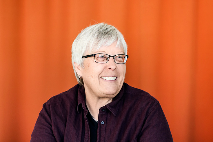 Kvinna med kort, grått hår och glasögon. Hon sitter framför en orange bakgrund och ler.