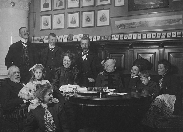 Äldre svartvitt familjeporträtt visar en stor familj med barn och vuxna runt ett bord.