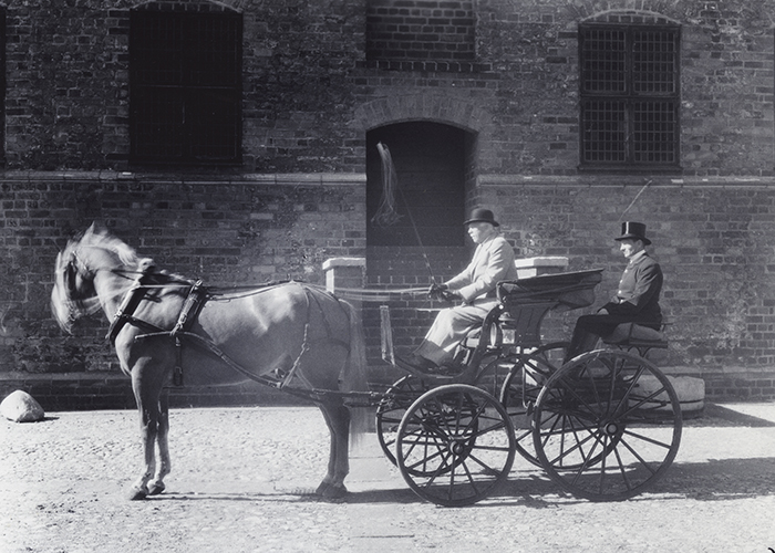 Äldre svartvitt fotografi där två män sitter i en öppen vagn bakom en häst