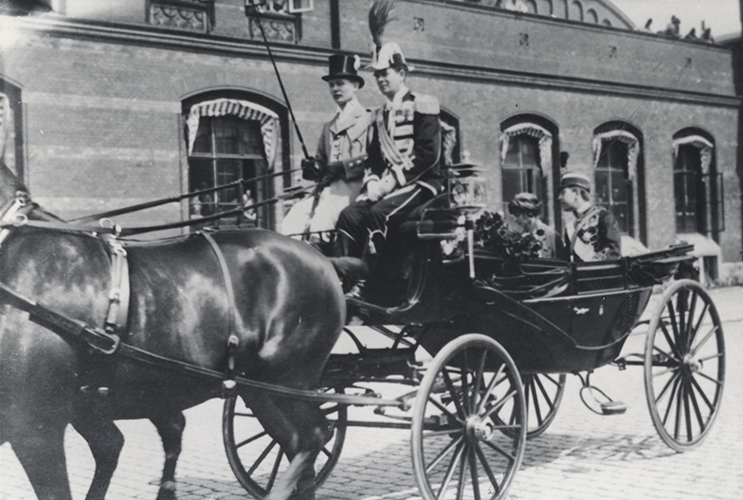En öppen fyrhjulig
vagn, med två hästar framför, kör längs en gata i Malmö. På kuskbocken sitter
en livréklädd kusk med hög hatt. Bredvid kusken sitter en uniformsklädd kunglig
betjänt. I vagnen sitter bl.a. kronprins Gustaf Adolf. Vagnen är landshövding
De la Gardies landå med kusken Gösta Åkerman. Fotot är taget i samband med
kronprinsens besök i Malmö 1926.  Foto:
Okänd fotograf / Malmö Museer.