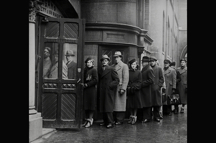 Män och kvinnor köar utanför entrén, de flesta i hatt och lång rock. 