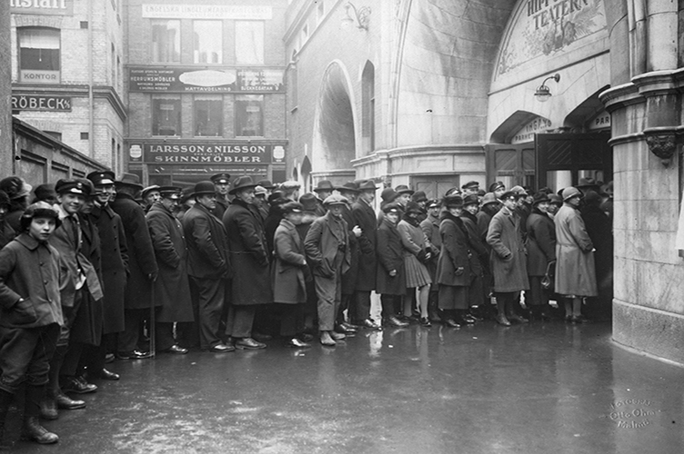 Äldre fotografi på många människor som står i kö till teatern, de allra flesta har rock och hatt.