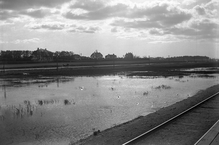 Svartvit äldre bild som visar ett tågspår jämte en damm och i bakgrunden några hus och träd.