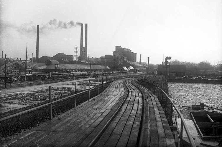 Svartvit äldre bild av cementfabrikens exteriör med järnvägsspår i förgrunden.
