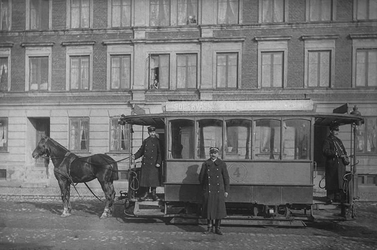 Svartvit äldre bild som visar en spårvagn dragen av en häst. I bakgrunden syns en äldre byggnad. På spårvagnen står män i konduktörsrockar.