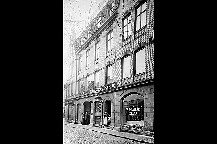 Äldre foto i svartvitt på ett av husen, byggnaden är 3 våningar hög med vindsfönster. Skyltfönstren vittnar om att här har bedrivits affärsverksamhet i bottenvåningen.