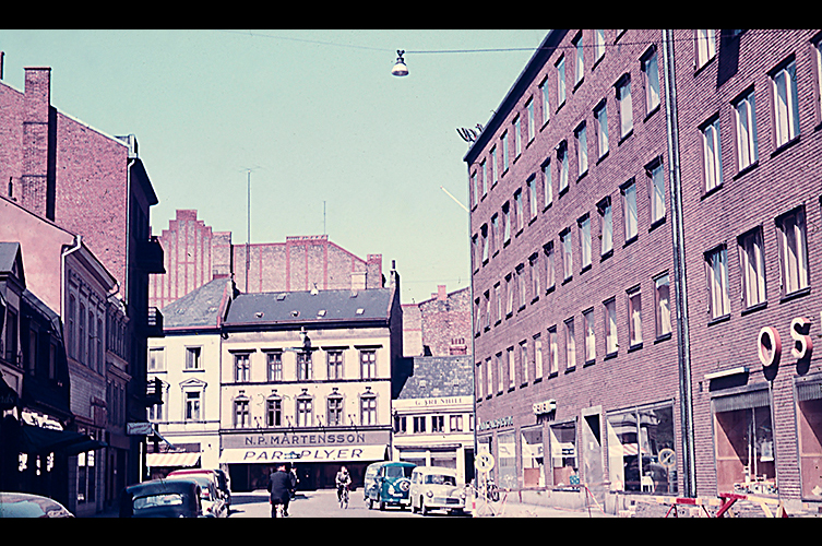 Äldre bild i färg som visar ett av de små husen på Djäknegatan. Runt om finns stora modernare byggnader. På huset finns en skylt där det står "Paraplyer".