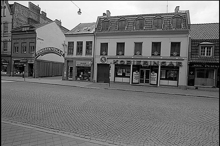 Äldre fotografi på hus vi Djäknegatan, i botten av huset ligger en butik och bredvid butiken ligger Pingstkyrkan. En bred gata med kullersten. 
