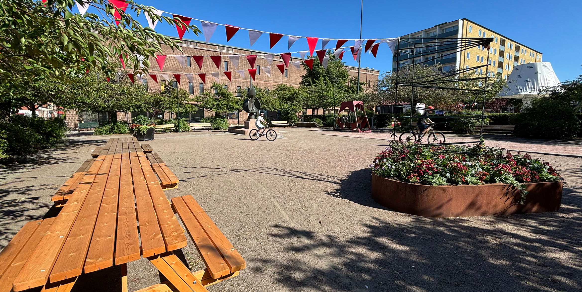 Två barn cyklar över ett torg där man även ser ett långbord i trä och girlanger från träd.