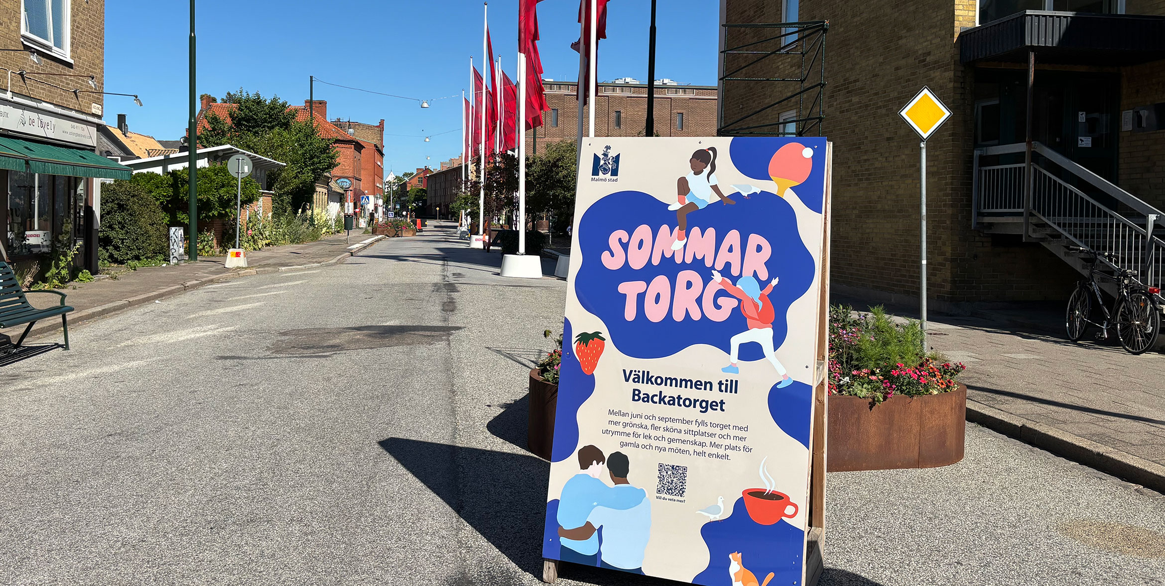 En skylt på en gata där det står sommartorg. Röda flaggor syns i bakgrunden.