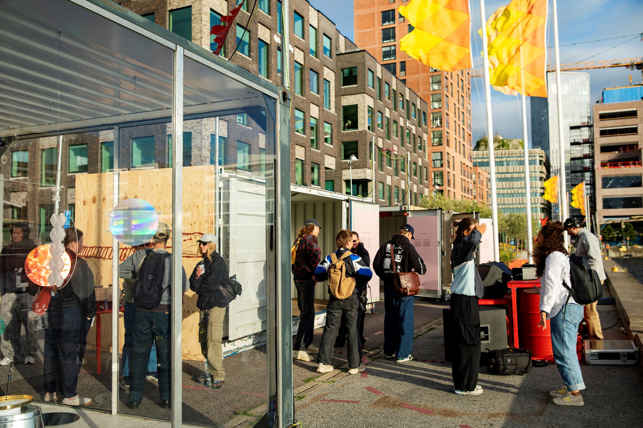 Besökare på vernissage den 3 juli. Folk minglar på en kaj vid en container och en glasbur. I bakgrunden flaggor som vajar.