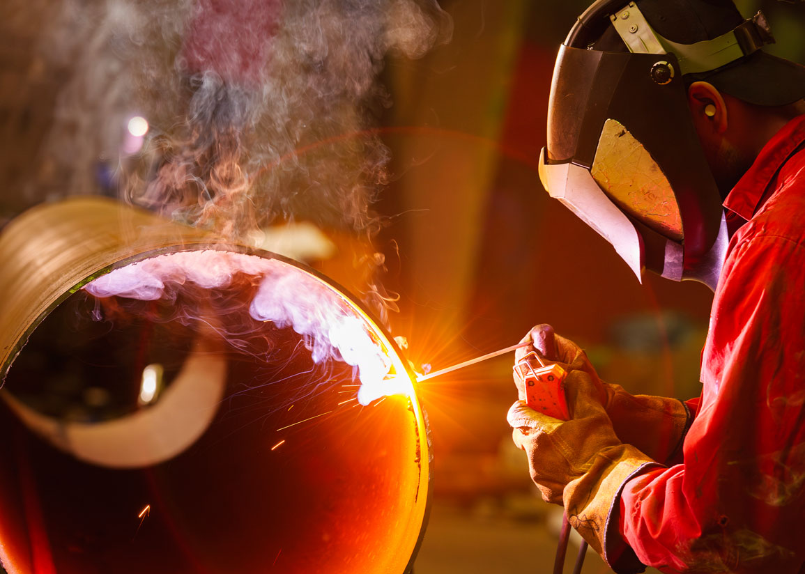 En rödklädd person med mask och handskar utför svetsning av metallrör, med kraftig ljuslåga och rök.