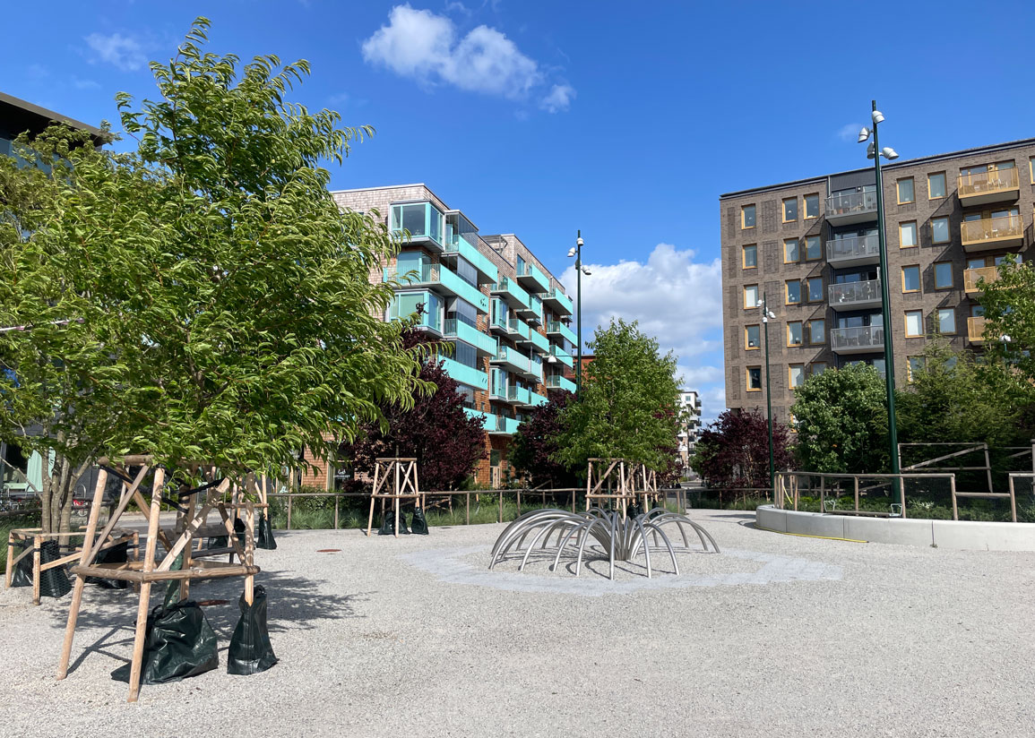 Nyanlagt torg i Limhamns hamnområde: Fendertorget med grusyta, träd och spindellik skulptur av metall. Bostadskvarter i bakgrunden en sommardag med blå himmel och lätta moln.