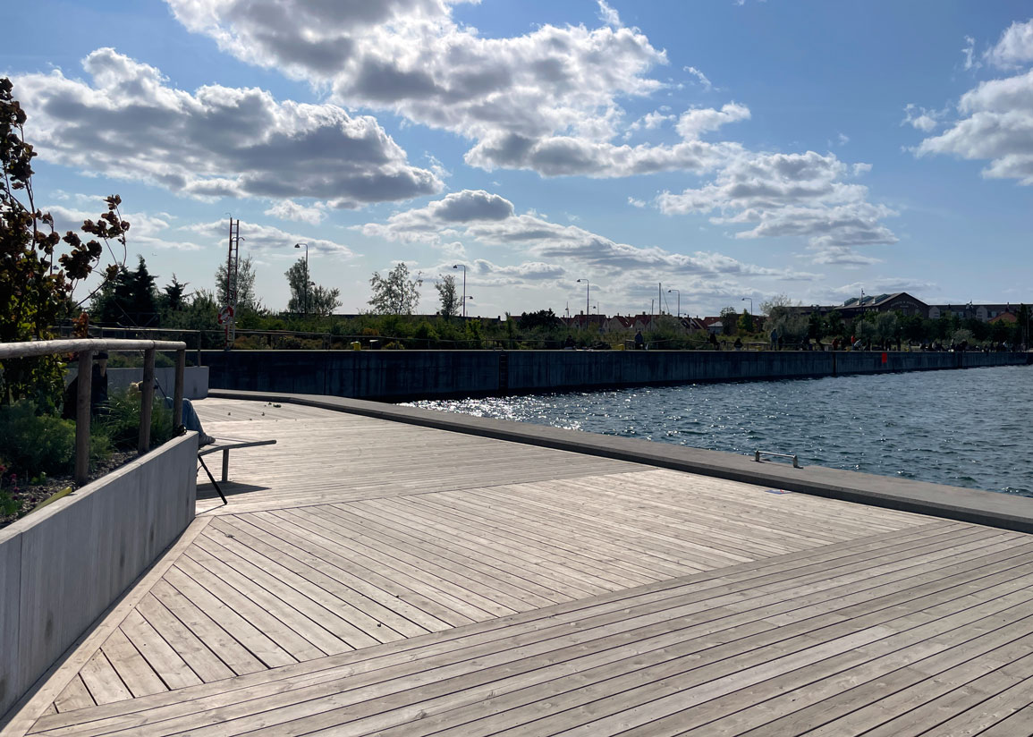 Cementkajen mot Övägen, med trädäck och sittplatser. Starkt solljus och stråk av moln på en blå himmel.