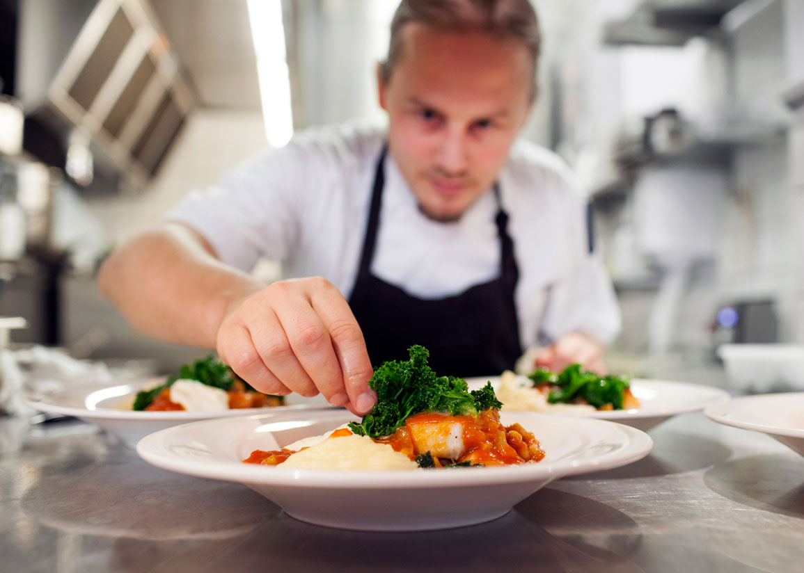 En man i ett restaurangkök lägger sista touchen på en maträtt i djup tallrik innan servering.