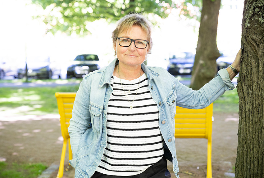 Marie står lutad mot ett träd med en av Malmö stads gula bänkar i bakgrunden.