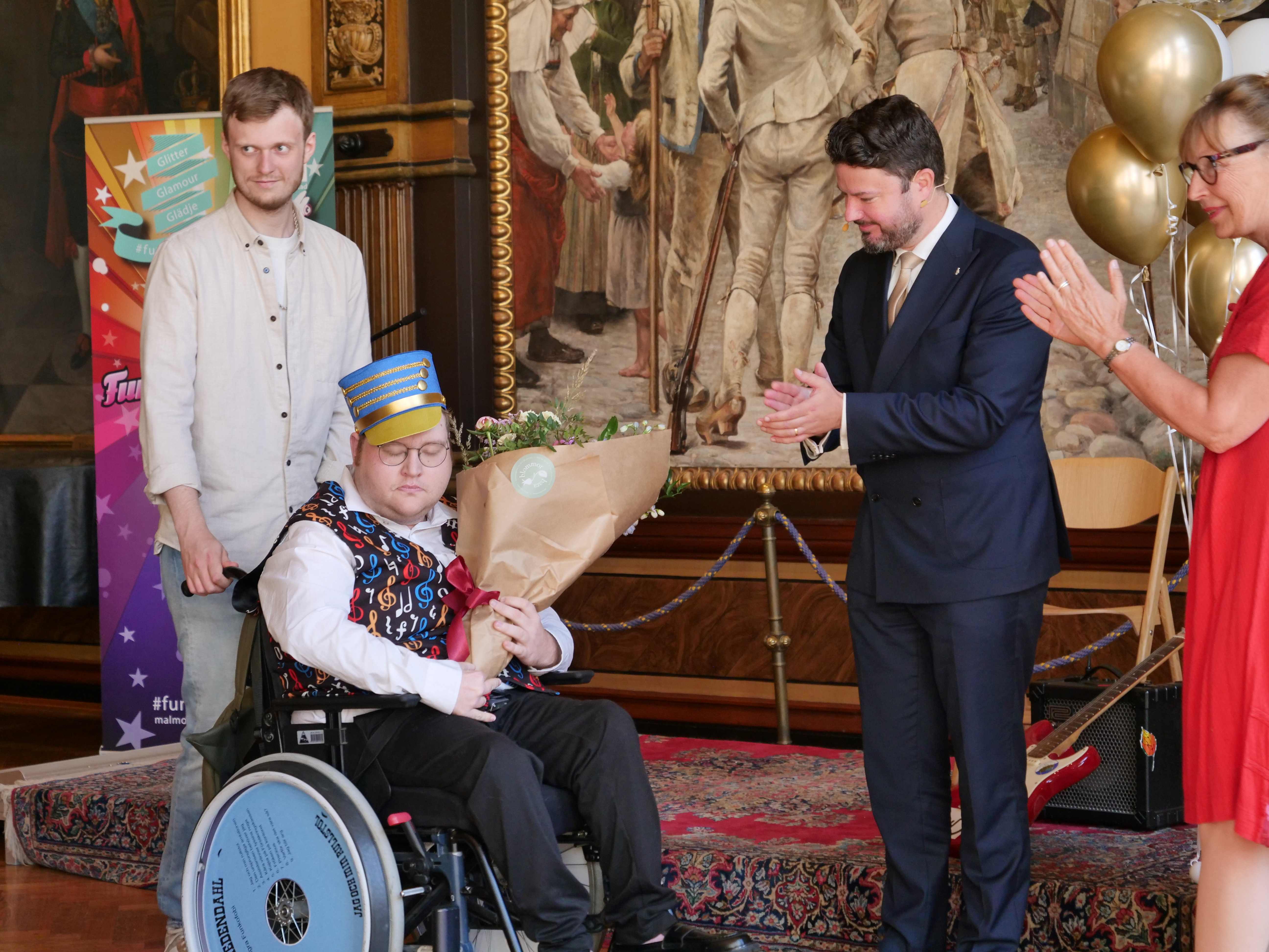William håller blommor i sin famn. Framför honom står Roko och Carina och applåderar. Bakom William står hans personliga assistent.