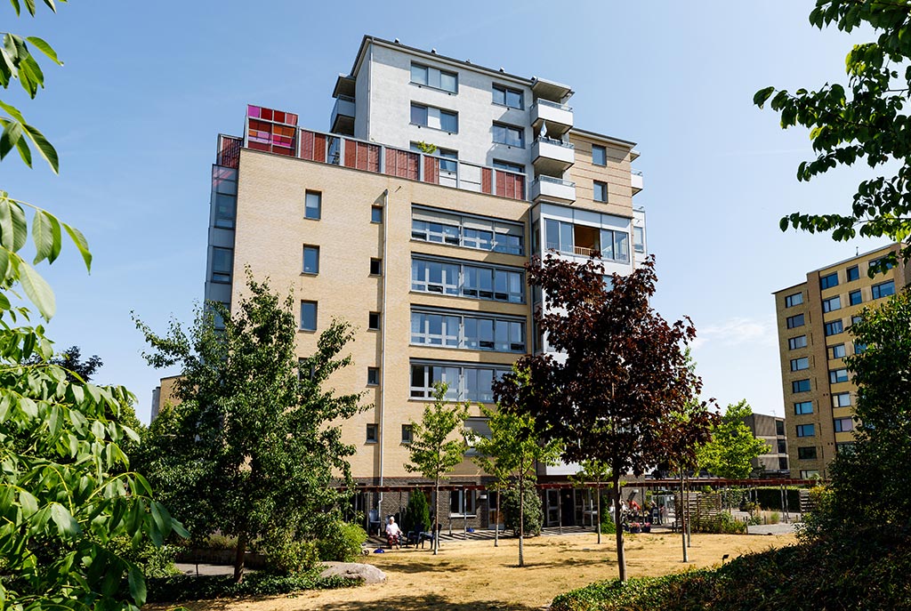 Huset är högt och man ser en färgglad terrass. På marken
framför huset finns en park med flertalet träd.