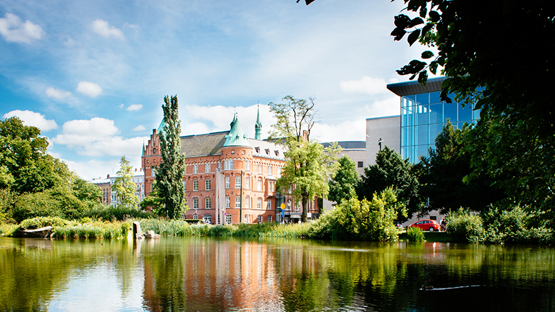 Stadsbiblioteket speglar sig i dammen i Slottsparken en solig sommardag, omgivet av grönska och blå himmel.