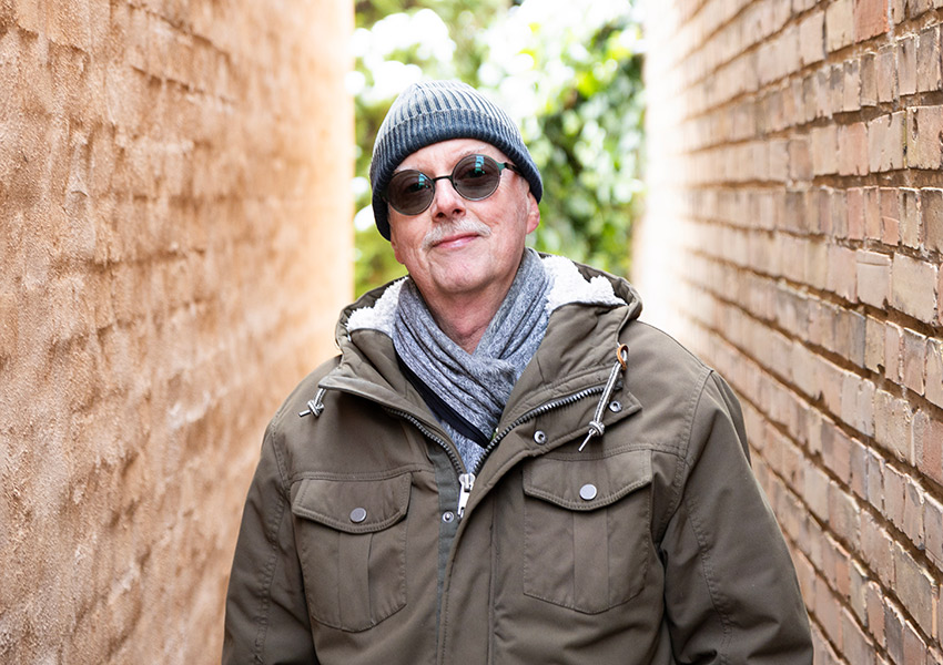 Claes Gylling har en mörkgrön jacka, grå sjal och mössa och runda solglasögon och står mellan två husväggar.