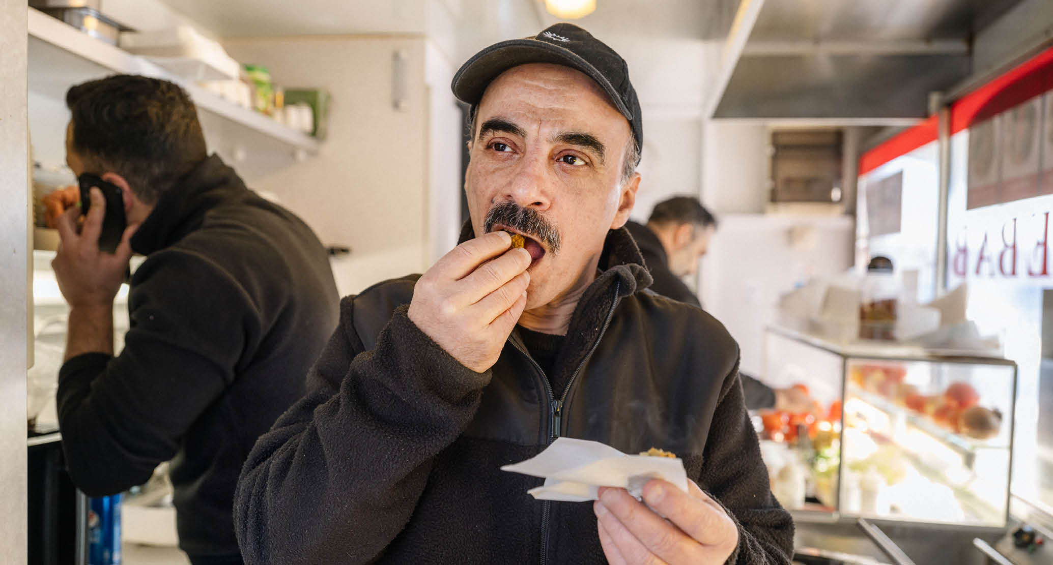 En man med mustasch i svart fleecejacka och keps äter en falafel.
I bakgrunden syns köket i en falafelvagn och två personer som arbetar. 