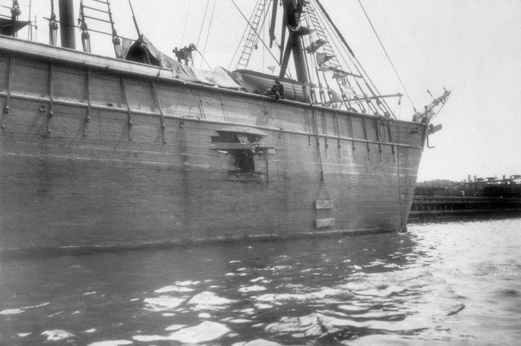 Äldre svart-vitt fotografi föreställande en båd. På båtens sidan syns ett hål orsakat av en explosion. 