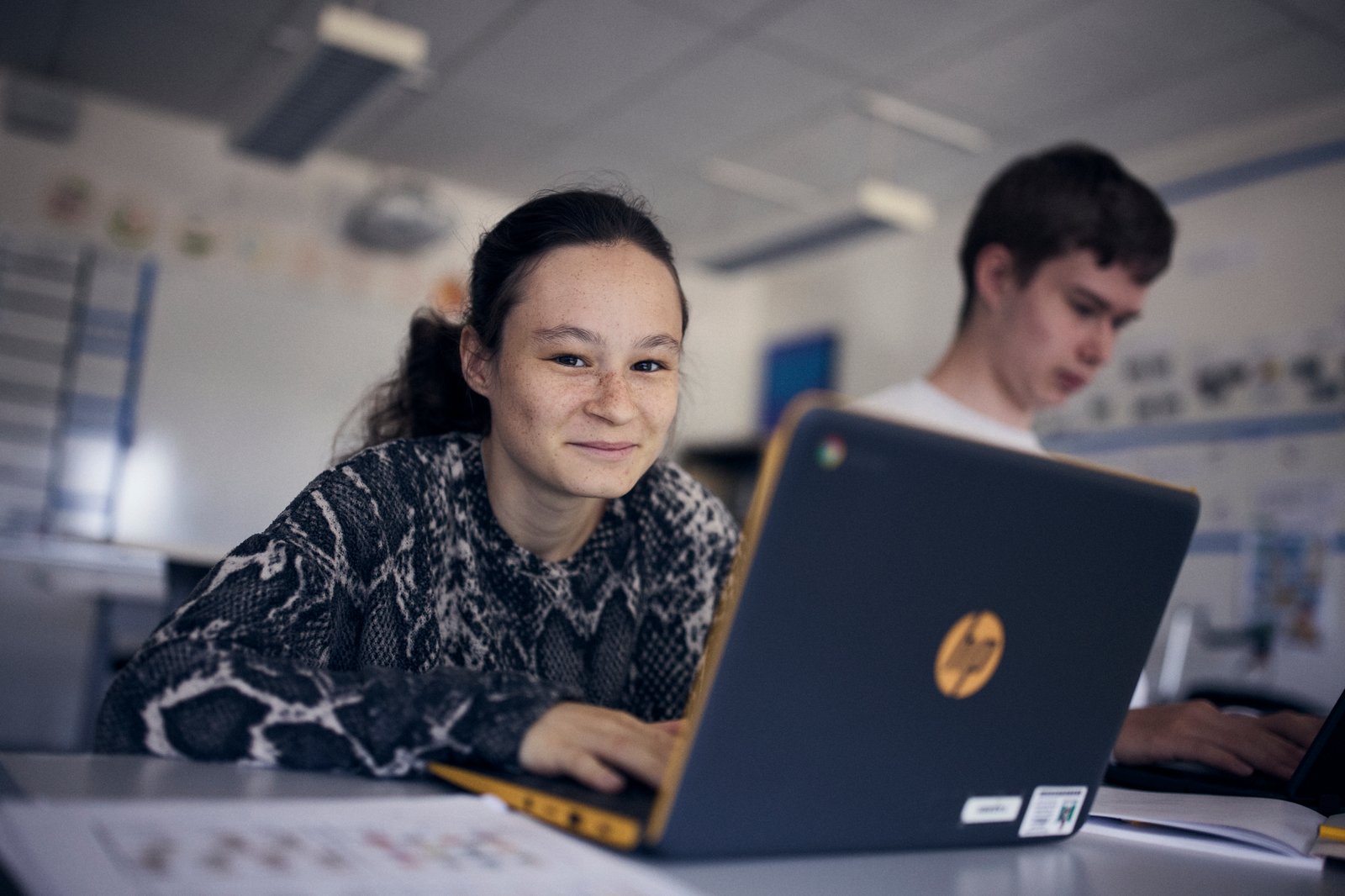 En elev sitter framför datorn och ler in i kameran och i bakgrunden syns en annan elev. 