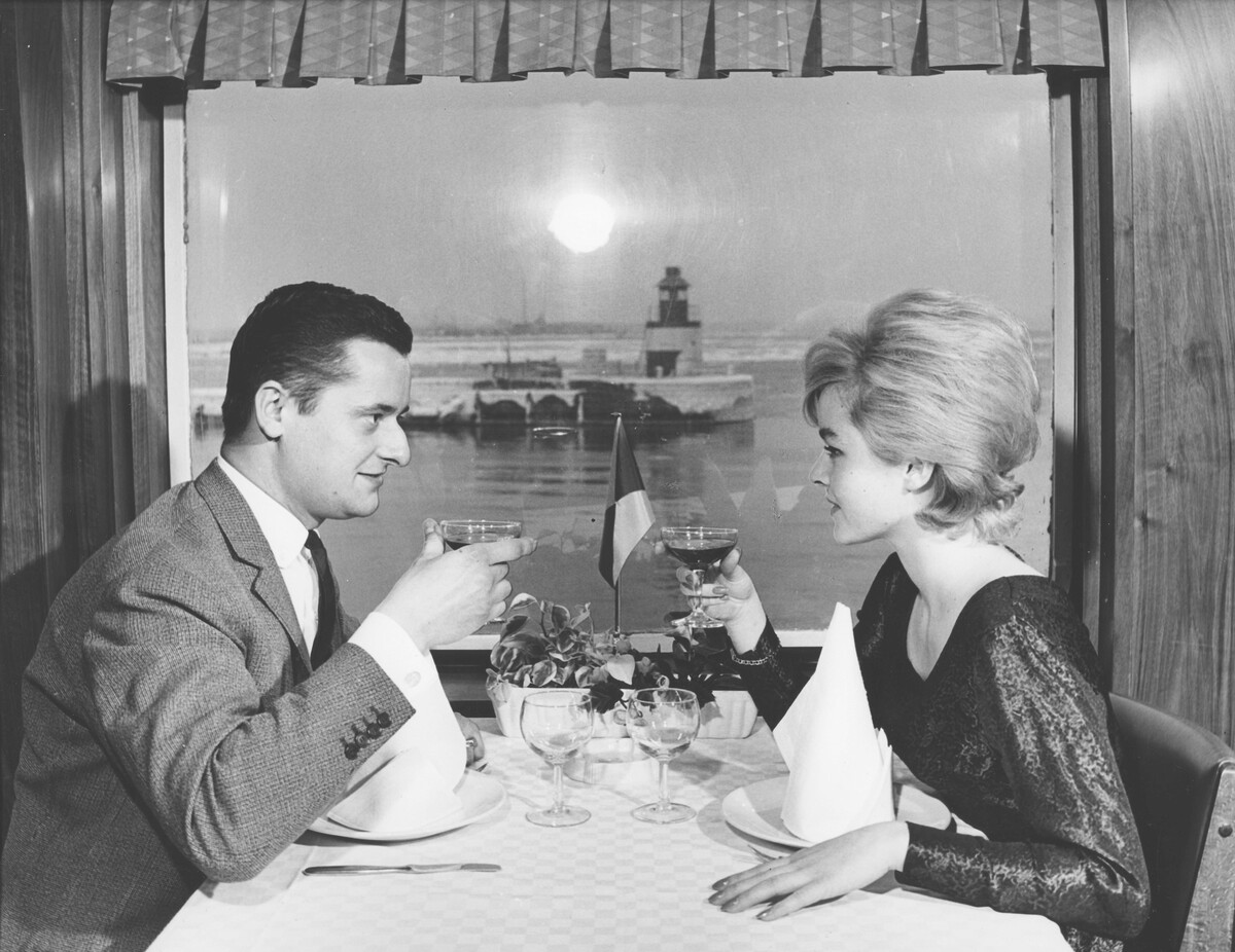Äldre svartvitt fotografi. Två personer mittemot varandra vid bord på färja.