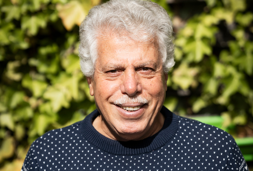 Mahmod ler stort. Han har lockigt grått hår och mustasch och är klädd i en blå stickad tröja.