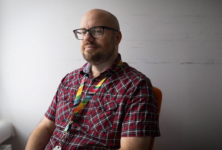 Tomas har svartbågade glasögon och skägg. Han har på sig en kortärmad rutig skjorta.