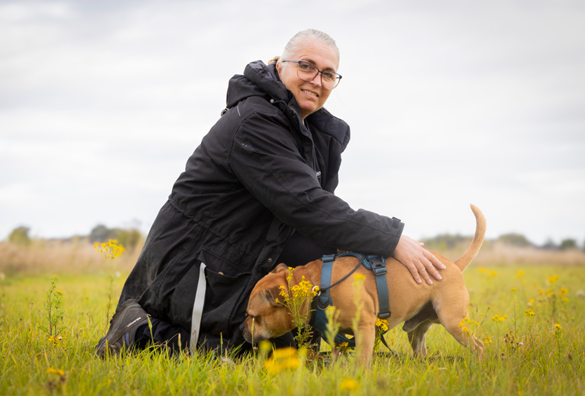 Anette har grått hår i hästsvans och svartbågade glasögon. Hunden Mamba luktar i fickan på hennes svarta jacka.
