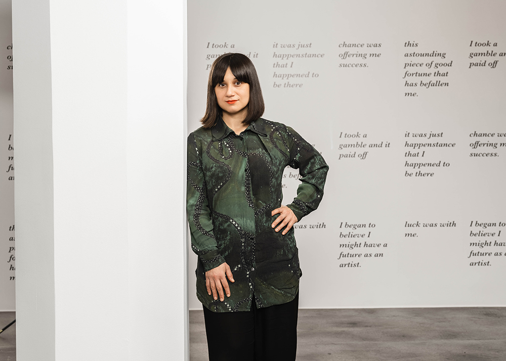 Porträttbild av Meriç Algün som står i grön skjorta framför en konstinstallation. Installationen är en vit vägg i ett ljust rum med citat skrivna i kursiv text.