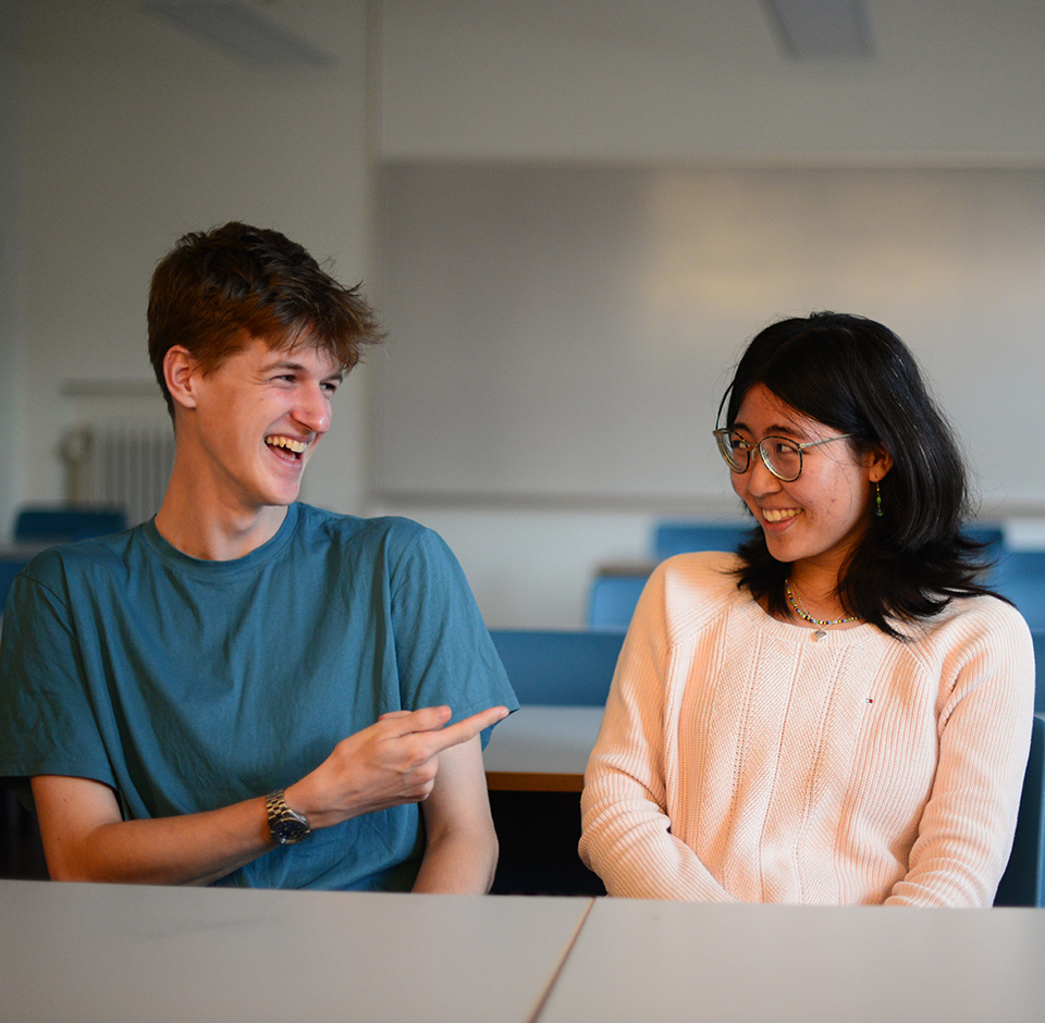 Två elever sitter i ett klassrum. De tittar på varandra och skrattar.