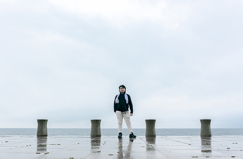 Man med svart tröja, vit scarf och beige byxor syns på håll, med hav och himmel i bakgrunden