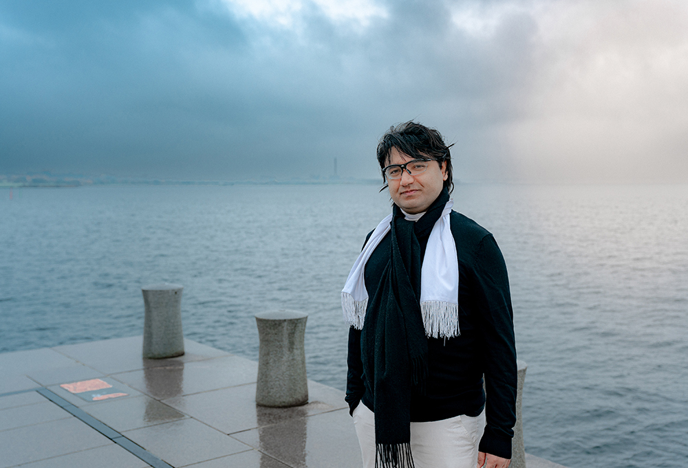 Chia småler mot kameran. Han har svart hår, glasögon, en svart tröja och en vit scarf. I bakgrunden syns hav, gråa moln och svagt solljus.
