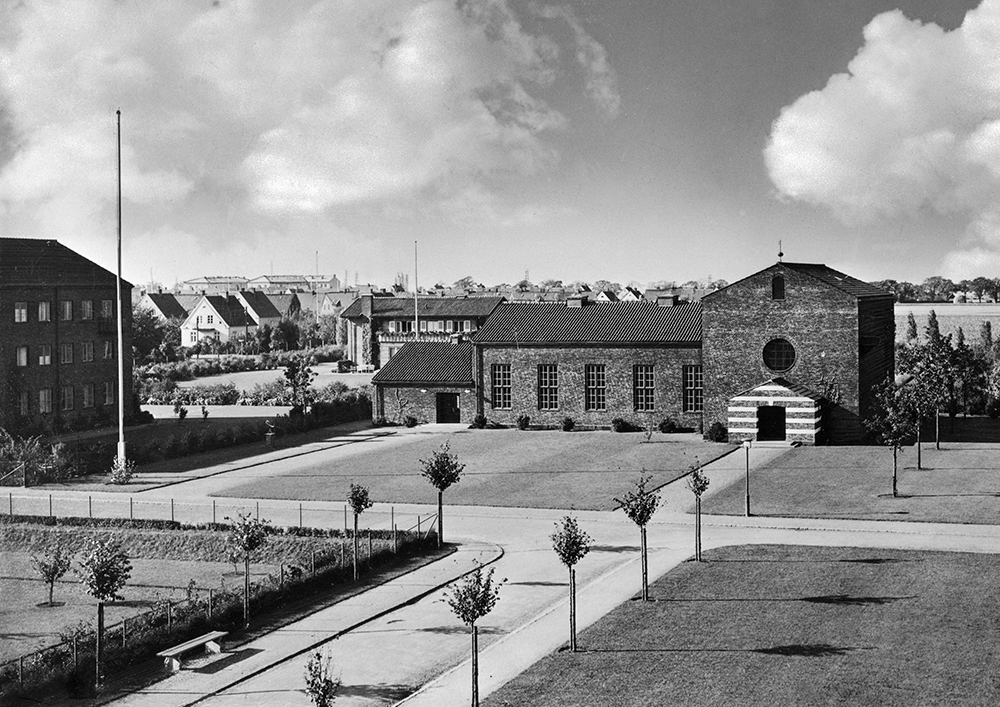 En kyrka i tegel med gräsmattor och en nyplanterad allé med träd. 
