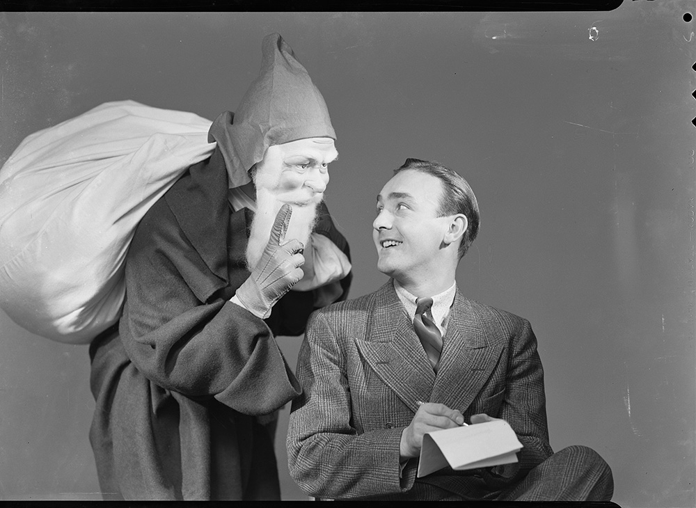 Äldre svartvitt fotografi. En i tomtemask står bakom en man klädd i kostym.
