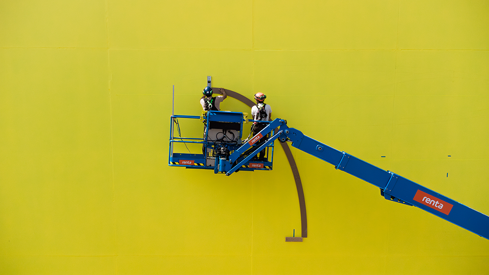 Luca Lee Frei skapar verket på plats. En lyftkran med två personer i syns mot en gull vägg.
